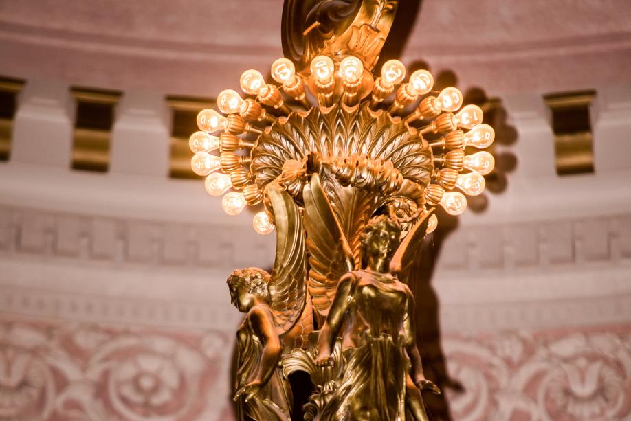 A close-up of the upper portion of large chandelier in the Legislative Building Rotunda. This section features two angels under a single row of small lightbulbs.