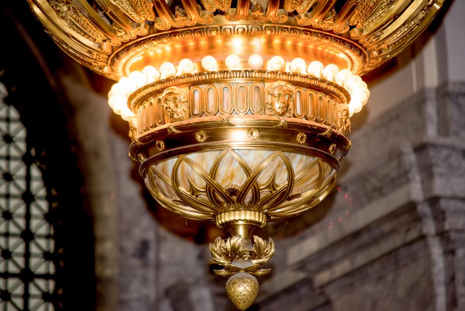 A close-up of the bottom portion of the large chandelier that hangs in the Legislative Building Rotunda. This section features small faces and flowers, white and gold marble, and a single row of small light bulbs.