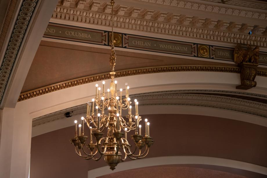 A polished bronze chandelier with lights that resemble candles. The chandelier hangs in the corner of a Legislative chamber room.