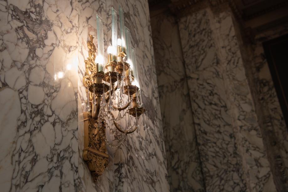 A polished bronze light fixture placed on a marble wall. The light fixture resembles a candle holder and has glass beads hanging from it.
