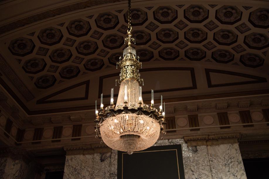 A polished bronze chandelier with thousands of glass beads on it hanging from a patterned ceiling. 