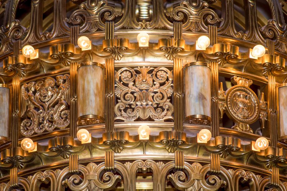 A close-up of the middle portion of the large chandelier that hangs in the Legislative Building Rotunda. This section features mythological creatures, an eagle, floral elements, and two rows of small light bulbs.
