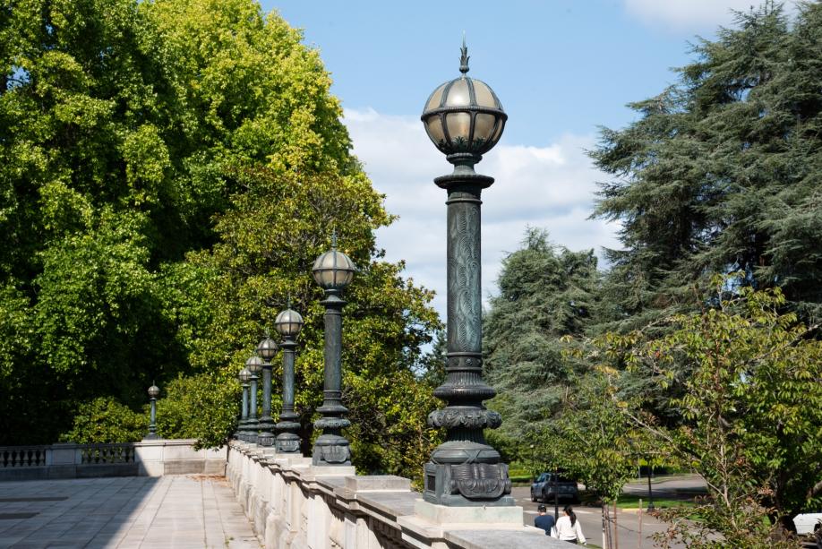 A row of vintage 1920s light posts standing on top of a sandstone railing. Large green trees and two people are in the background.