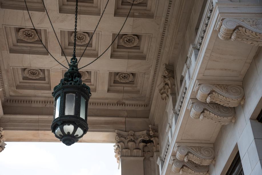 A large bronze lantern hanging from five chains above the south entrance of the Legislative Building. Intricate designs are carved in sandstone walls and ceiling around the lantern.