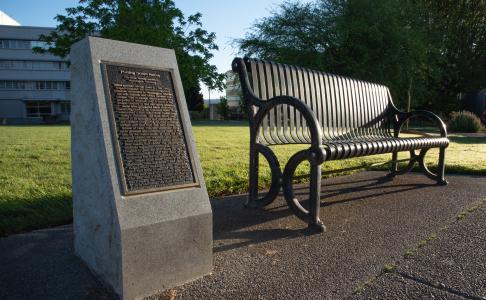 The George Bush Monument, a bronze plaque set in a granite slab, next to a metal park bench with two large trees and the General Administration building in the background.