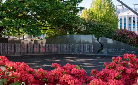 A patch of beautiful red flowers in the foreground surrounds the granite Vietnam Veterans Memorial, featuring small American flags hanging next to the names of Washington state residents killed or missing during the Vietnam War, which is shaded by the trees behind it.