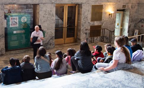 A tour guide smiling at a group of children sitting on marble steps inside the Legislative Building.