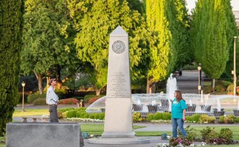 A man and a woman standing on both sides of the Medal of Honor Memorial, reading its inscriptions, with the Tivoli Fountain and green trees in the background.