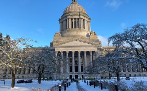 A snow-covered walkway and lawn facing the south side of the Legislative Building. A grounds crew member operates a small snowplow in the distance.