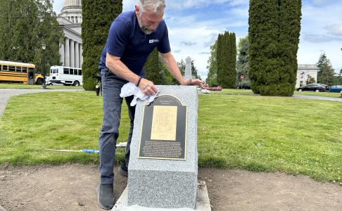 DeWayne Steinman, a DES Buildings & Grounds employee, is shown polishing the new bronze plaque that rests atop a granite base on the Washington State Capitol Campus with trees, bushes, grass, and blue sky in the background.