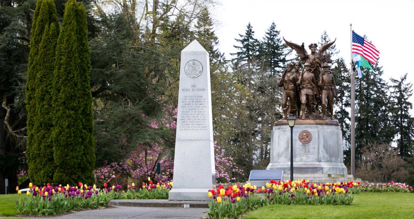 An outdoor scene with a stone pathway lined with tulips leading to the Medal of Honor Memorial, with the Winged Victory Monument, trees, and the American flag in the background.