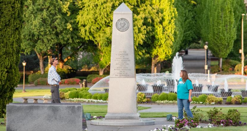 A man and a woman standing on both sides of the Medal of Honor Memorial, reading its inscriptions, with the Tivoli Fountain and green trees in the background.