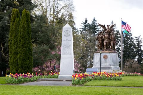 An outdoor scene with a stone pathway lined with tulips leading to the Medal of Honor Memorial, with the Winged Victory Monument, trees, and the American flag in the background.