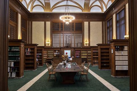 The Washington Law Library lobby, which has a dark green carpet and a brown wooden table in the middle of the room, with brown wooden bookshelves on both sides of the table. A large chandelier hangs above the front desk, where a person is talking to two clerks. 