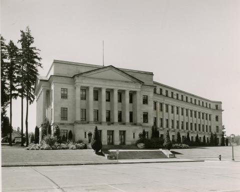 A black and white image of the front side of Cherberg Building from across the street. Three large evergreen trees stand to the left of the building, and many smaller trees and plants line the exterior of the building.