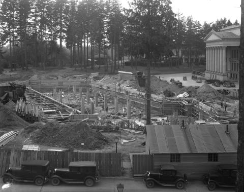 A black and white image of a construction site lined with a wooden fence and black cars. The south side of the Legislative Building, the Governor's Mansion, and trees are visible in the background.