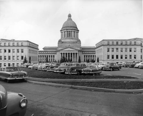 A black and white image of the John L. O'Brien and John A. Cherberg Building framing the Legislative Building. A full parking lot is in the foreground, and a plaza with small evergreen trees is visible between the buildings.