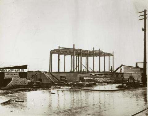 A black and white image of the Dolliver Building's construction site. Wooden framework stands on a concrete foundation covered in rainwater, with construction workers visible throughout the site.