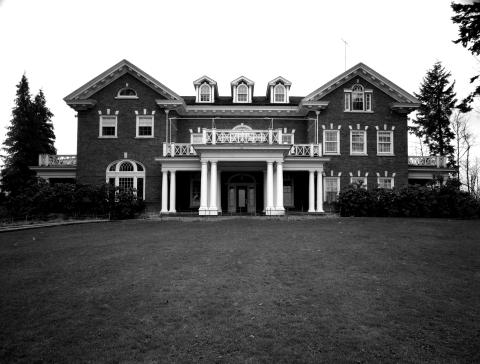 A black and white image of the front side of the Governor's Mansion. A lawn goes all the way up to the front porch, as there is no driveway yet, and bushes line the entire front side of the building.