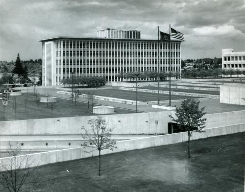 A black and white image of the front of the Highways-Licenses Building taken from across the East Capitol Campus Plaza. The building stands tall above groves of trees and a large, grassy plaza with the Washington State Flag and American Flag waving in the wind.