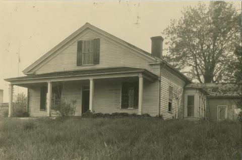 A black and white image of Isaac Stevens' abandoned house, which is white and overgrown with plants. The house sits on a small hill in front of a large tree.