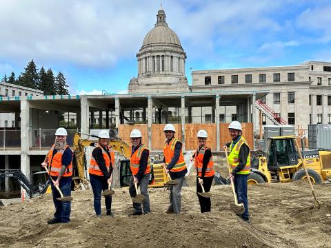 Six individuals from the Legislative Campus Modernization (LCM) Project team with reflective vests and hardhats are smiling while shoveling dirt at the Joel M. Pritchard construction site. Part of the Prichard, Legislative, and Cherberg buildings are visible in the background.