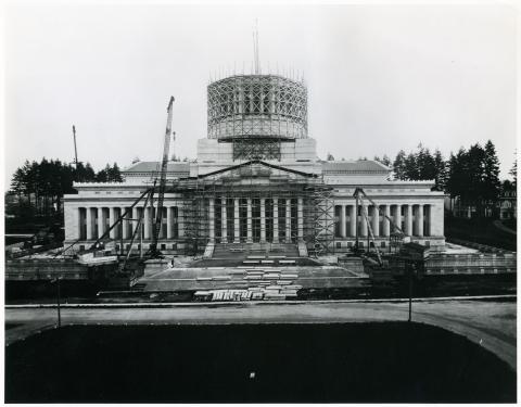 A black and white image of the front side of the Legislative Building, showing the building before its dome was built. The building is surrounded by a crane, scaffolding, stone slabs, and other construction materials.