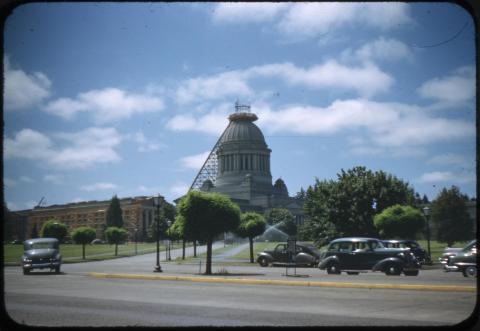 A wide shot of wooden scaffolding climbing up to the very the Legislative Building dome, which is being repaired after an earthquake. Parts of the West Capitol Campus lawn and the Insurance Building are visible, with multiple trees and cars in the foreground.