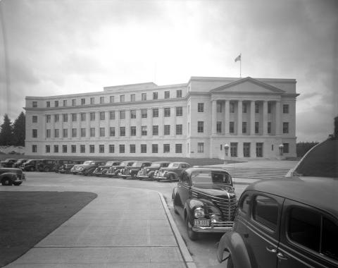 A black and white image of the front side of the John L. O'Brien Building from down the street. Black cars fill the parking lot in front of the building, and some trees are visible behind the building.