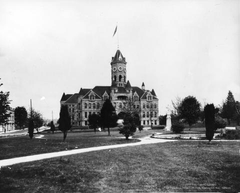 A black and white image of the front side of the Old Capitol Building taken from Sylvester Park across the street. The building has a tall tower in the middle of it, where a flag waves in the wind.