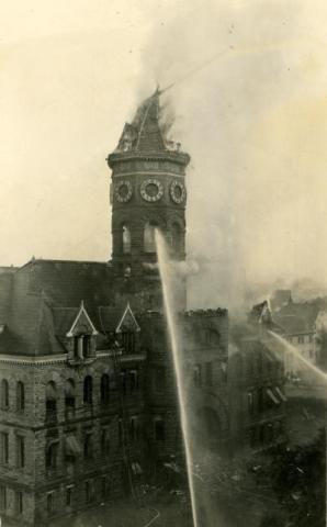 A black and white image of the front of the Old Capitol Building in flames. Streams of water coming from firehoses are spraying the building's tower and front windows, and a ladder leads firemen to the rooftop.