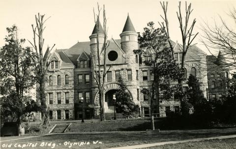 A black and white image of the front of the Old Capitol Building taken from Sylvester Park across the street. The tall tower that once stood at the top of the building is now gone. Writing at the bottom of the photo says "Old Capitol Bldg. - Olympia, Wn."