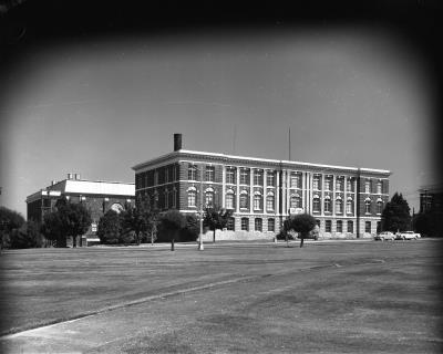 A black and white image of the old Olympia High School site taken from afar. The brick building is three stories tall and faces a street lined with trees. Two white cars are parked in front of the school.