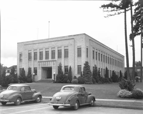 A black and white image of the original Irving R. Newhouse Building taken from across the street. Two gray cars are parked in front of the building, which is surrounded by small evergreen trees with three larger evergreen trees to its right.