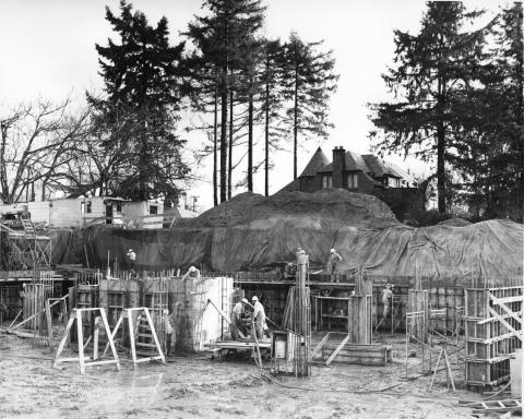 A black and white image of a construction crew working on an excavated site. Two houses and large evergreen trees are visible in the background.