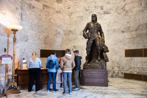 The Marcus Whitman statue standing next to the front desk at the north entrance of the Legislative Building. A group of visitors are talking to a Visitor Services representative at the front desk.