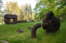 A wide shot of the Boiler Works statue, made of two large steel structures and two chained steel bulbs, spanning across a small grassy hill with trees in the background. 