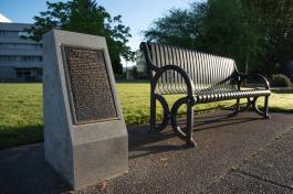 The George Bush Monument, a bronze plaque set in a granite slab, next to a metal park bench with two large trees and the General Administration building in the background.