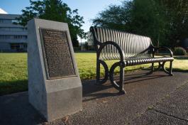 The George Bush Monument, a bronze plaque set in a granite slab, next to a metal park bench with two large trees and the General Administration building in the background.