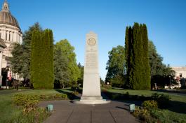 The Medal of Honor Memorial, a white granite obelisk, stands on a stone pathway with trees and the Legislative Building in the background.