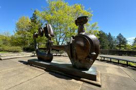 The Shaman Statue, a large bronze structure surrounded by metal benches, reflects the sunshine with tall trees in the background.