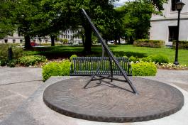 An image of the Territorial Sundial, made of bronze rods set on top of a circular brass platform, with a metal park bench and cherry blossoms in the background.