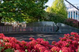 A patch of beautiful red flowers in the foreground surrounds the granite Vietnam Veterans Memorial, featuring small American flags hanging next to the names of Washington state residents killed or missing during the Vietnam War, which is shaded by the trees behind it.