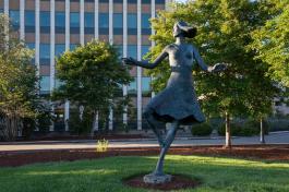 The bronze Woman Dancing Statue, depicting a woman twirling, stands tall in the grass with trees and the Highways and Licenses building in the background.