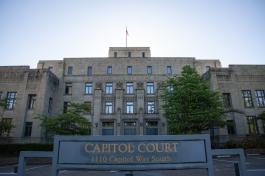 A wide shot of the Capitol Court Building, which is a four-story stone building with large blue doors and an American flag waving on the roof. The words "Thurston County Court House" are inscribed in the stone near the top of the building, and there is a blue sign with gold lettering in the foreground that says, "Capitol Court, 1110 Capitol Way South."