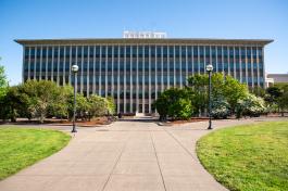 A wide shot of the front of the Highways-Licenses Building, which is five stories tall and made of white and brown stone. A concrete pathway lined with grass, trees, and light posts leads to the building's entrance.