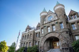 The main entrance of the Old Capitol Building, which is made of large, grayish-brown stone bricks and has two towers with teal cone-shaped roofs. A flagpole, a statue, and lots of trees are standing around the entrance.