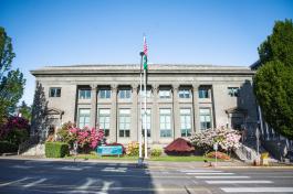 The front of the Dolliver Building, which is a rectangular stone building with Greek-style columns and large windows. Flagpoles waving the Washington State Flag and American Flag, along with flowering trees and bushes, stand in front of the building.