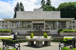 A view of the Joel Pritchard State Library's front side, which is mainly made of white stone with large glass walls. A fountain, a sundial, four metal park benches, and various green bushes are in front of the building.