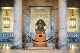 A large bronze statue of President George Washington's head, which sits on a wooden pedestal behind a rope barrier. Two marble columns stand on each side of the statue, and two large bronze chandeliers light up the area.